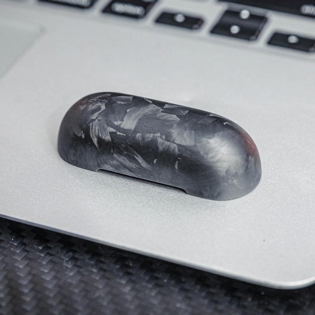 Close-up of the top cover of a forged carbon fiber AirPods case showcasing the unique marbled texture, placed on a silver laptop surface with a carbon fiber textured background and keyboard elements visible.