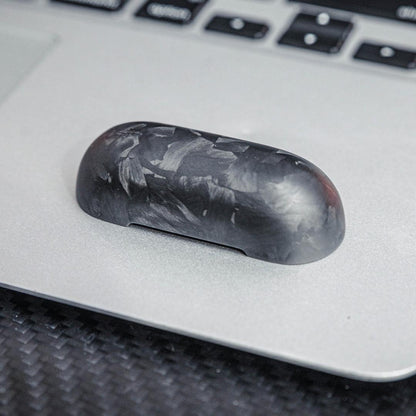Close-up of the top cover of a forged carbon fiber AirPods case showcasing the unique marbled texture, placed on a silver laptop surface with a carbon fiber textured background and keyboard elements visible.