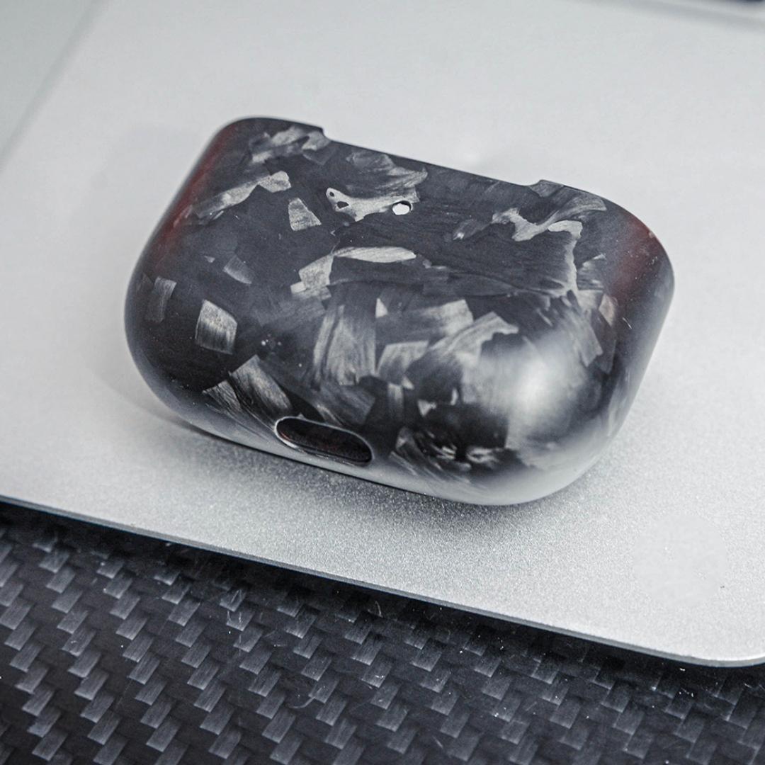 Close-up of the back side of the lower half of a forged carbon fiber AirPods case, showcasing its unique marbled texture, placed on a silver surface with a carbon fiber textured background.