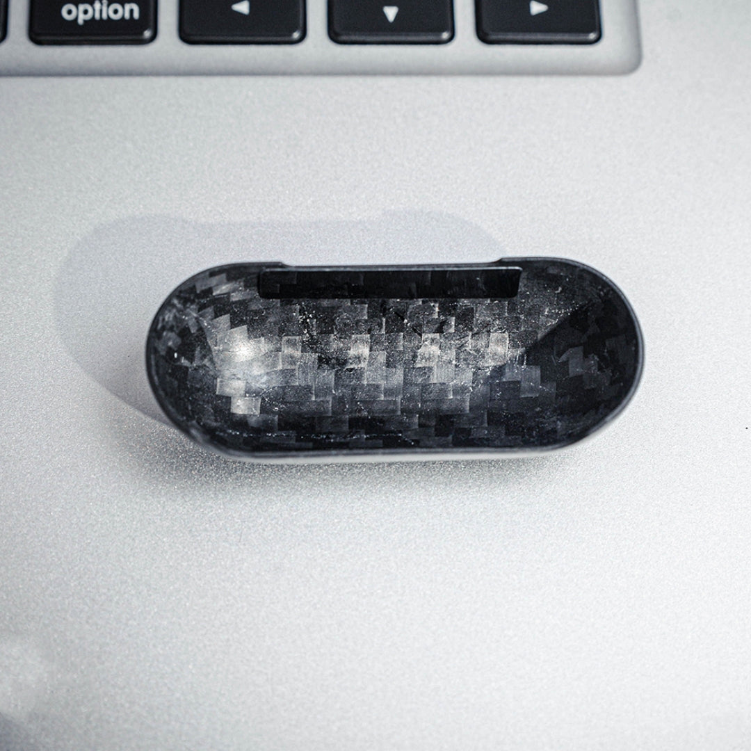 Close-up of the interior of the top half of a carbon fiber AirPods case, showcasing the distinct weave pattern, placed on a silver surface with a keyboard visible in the background.