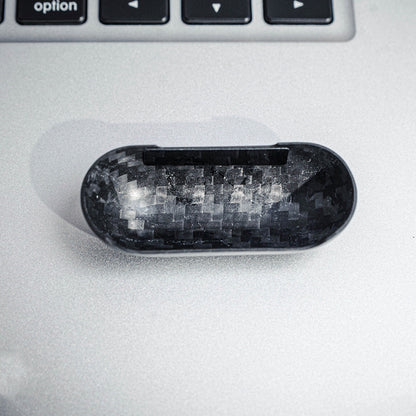 Close-up of the interior of the top half of a carbon fiber AirPods case, showcasing the distinct weave pattern, placed on a silver surface with a keyboard visible in the background.