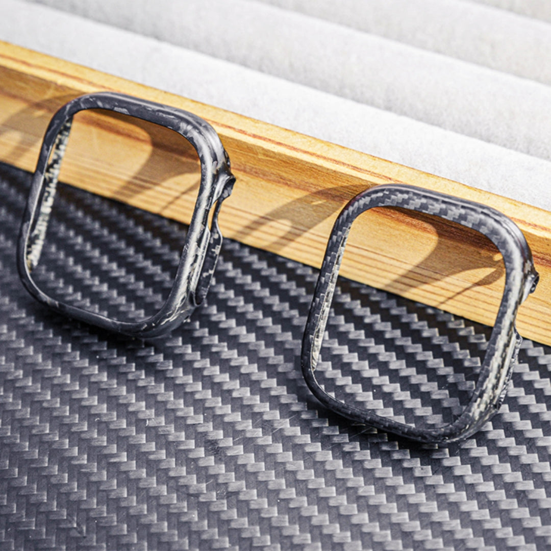 Close-up of two Apple Watch cases—one with 3K carbon fiber woven texture and the other with forged carbon fiber marbled texture—displayed on a carbon fiber-textured surface with wooden and soft-lined accents in the background.