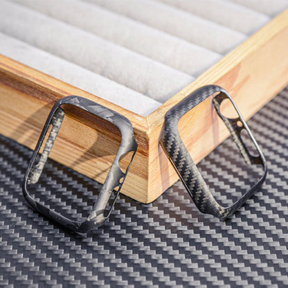 Close-up of two Apple Watch cases—one with 3K carbon fiber woven texture and the other with forged carbon fiber marbled texture—displayed on a carbon fiber-textured surface with wooden accents in the background.