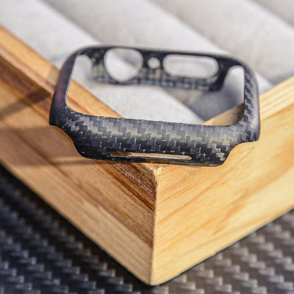 Close-up of a 3K carbon fiber Apple Watch case with a distinct woven texture, featuring precise cutouts, displayed on a wooden accent against a carbon fiber-textured surface.