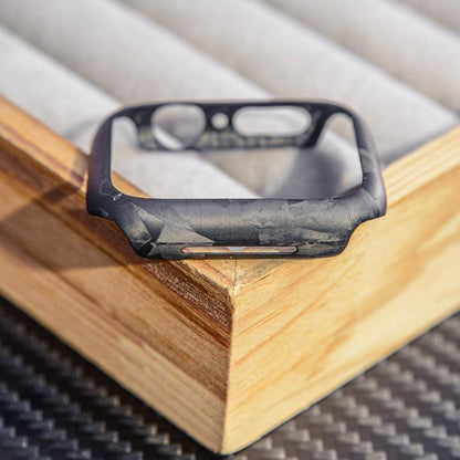 Close-up of a forged carbon fiber Apple Watch case with a distinct marbled texture, featuring precise cutouts, displayed on a wooden accent against a carbon fiber-textured surface.