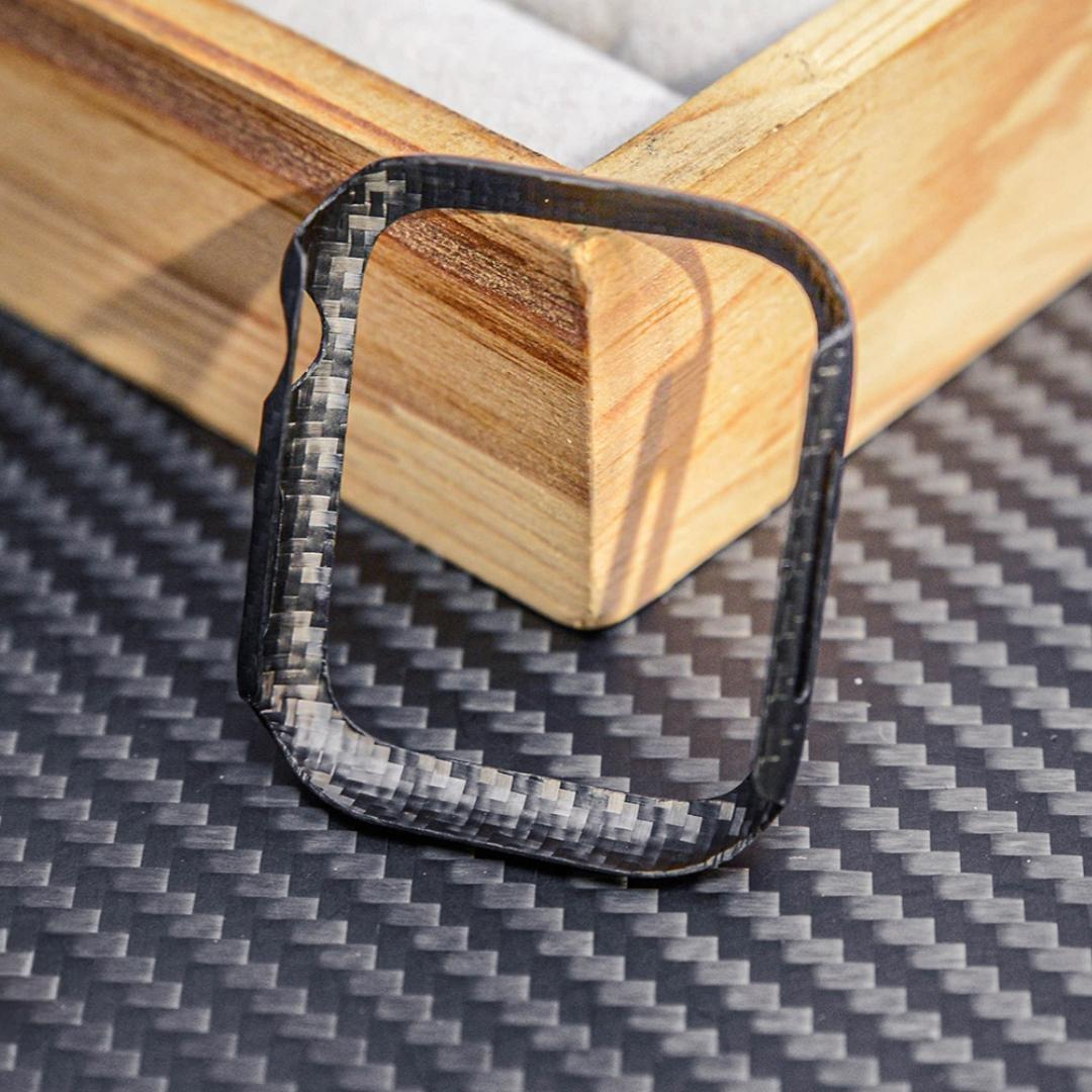 Close-up of a forged carbon fiber Apple Watch case with a distinct marbled texture, displayed on a carbon fiber-textured surface with wooden accents in the background.