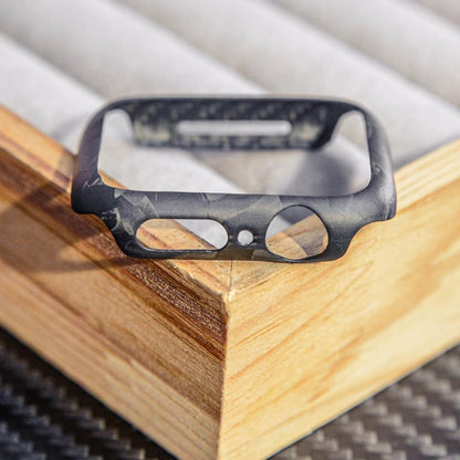 Close-up of a forged carbon fiber Apple Watch case with a distinct marbled texture, featuring precise cutouts, displayed on a wooden accent against a carbon fiber-textured surface.
