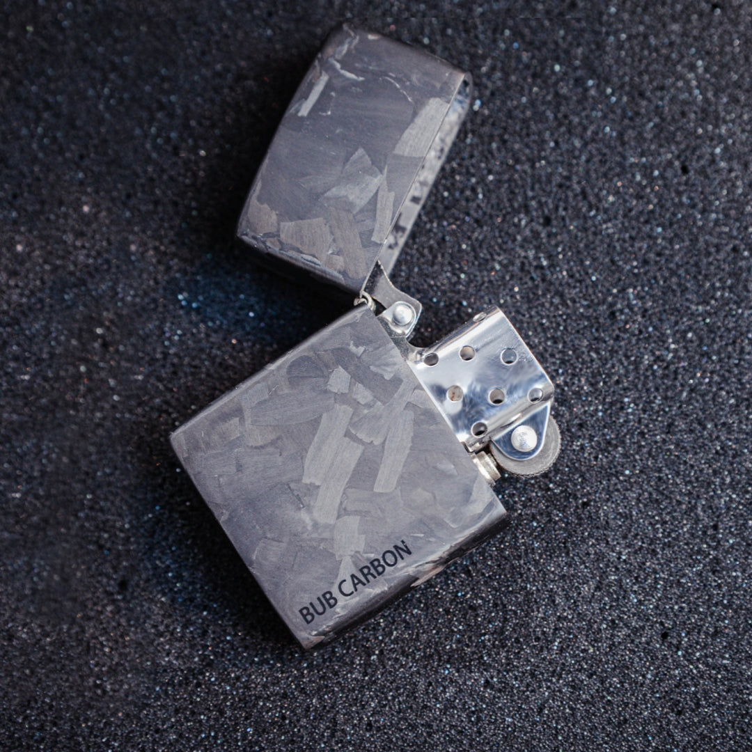 Close-up of an open Zippo lighter with a forged carbon case featuring the "BUB CARBON" branding, displayed on a textured dark surface.