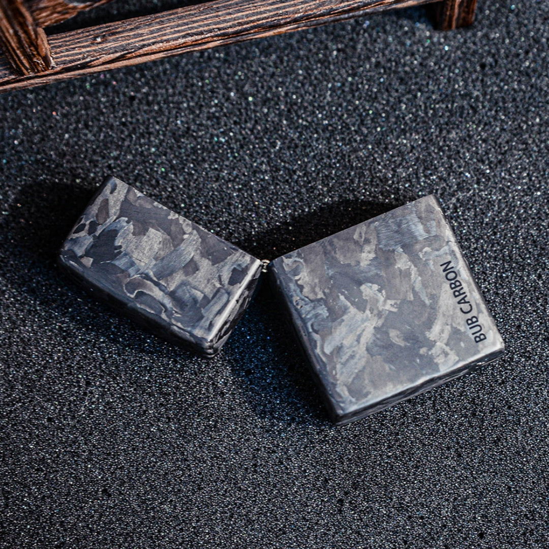 Close-up of an open forged carbon fiber Zippo lighter case with "BUB CARBON" branding, showcasing the unique marbled texture, placed on a textured dark glittery surface with a wooden structure in the background.