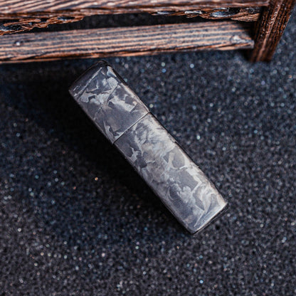 Close-up of a closed Zippo lighter case with forged carbon fiber construction, showcasing its unique marbled texture, placed on a textured dark glittery surface with a wooden structure in the background.