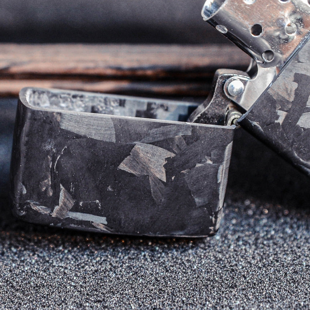 Close-up of an open Zippo lighter case with forged carbon fiber construction, showcasing the detailed marbled texture, placed on a textured dark surface with a wooden element in the background.