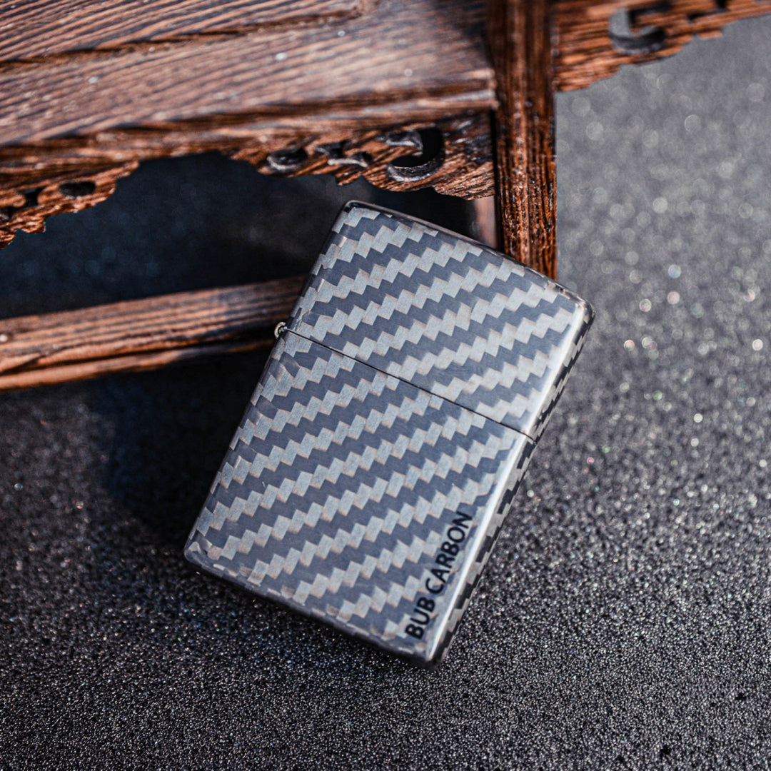 Close-up of a closed 3K carbon fiber Zippo lighter case showcasing the distinct weave pattern and "BUB CARBON" branding, placed on a dark glittery surface with a textured wooden structure in the background.