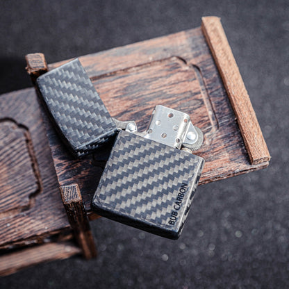 Close-up of an open 3K carbon fiber Zippo lighter case showcasing the distinct weave pattern and "BUB CARBON" branding, positioned on a textured wooden structure against a glittery background.