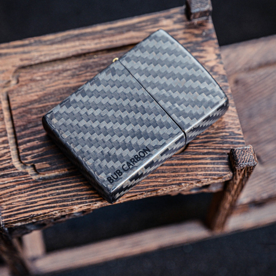 Close-up of a closed 3K carbon fiber Zippo lighter case showcasing the distinct weave pattern and "BUB CARBON" branding, placed on a textured wooden surface.