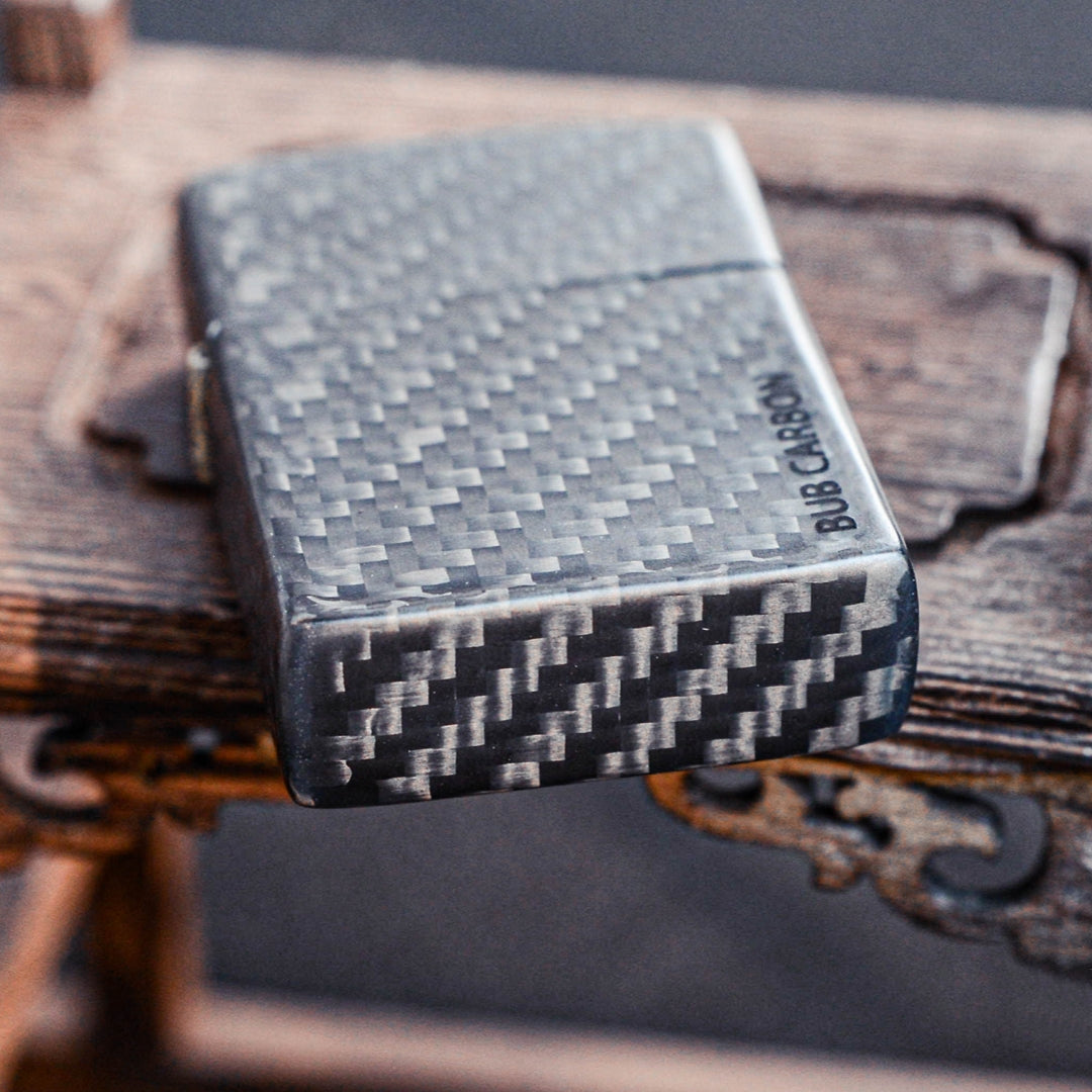 Close-up of a 3K carbon fiber Zippo lighter case showcasing the distinct weave pattern and "BUB CARBON" branding, placed on a textured wooden surface with carved details.