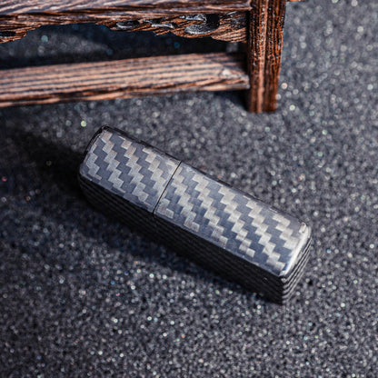Close-up of a closed 3K carbon fiber Zippo lighter case showcasing the distinct weave pattern, placed on a textured glittery dark surface with a carved wooden structure in the background.