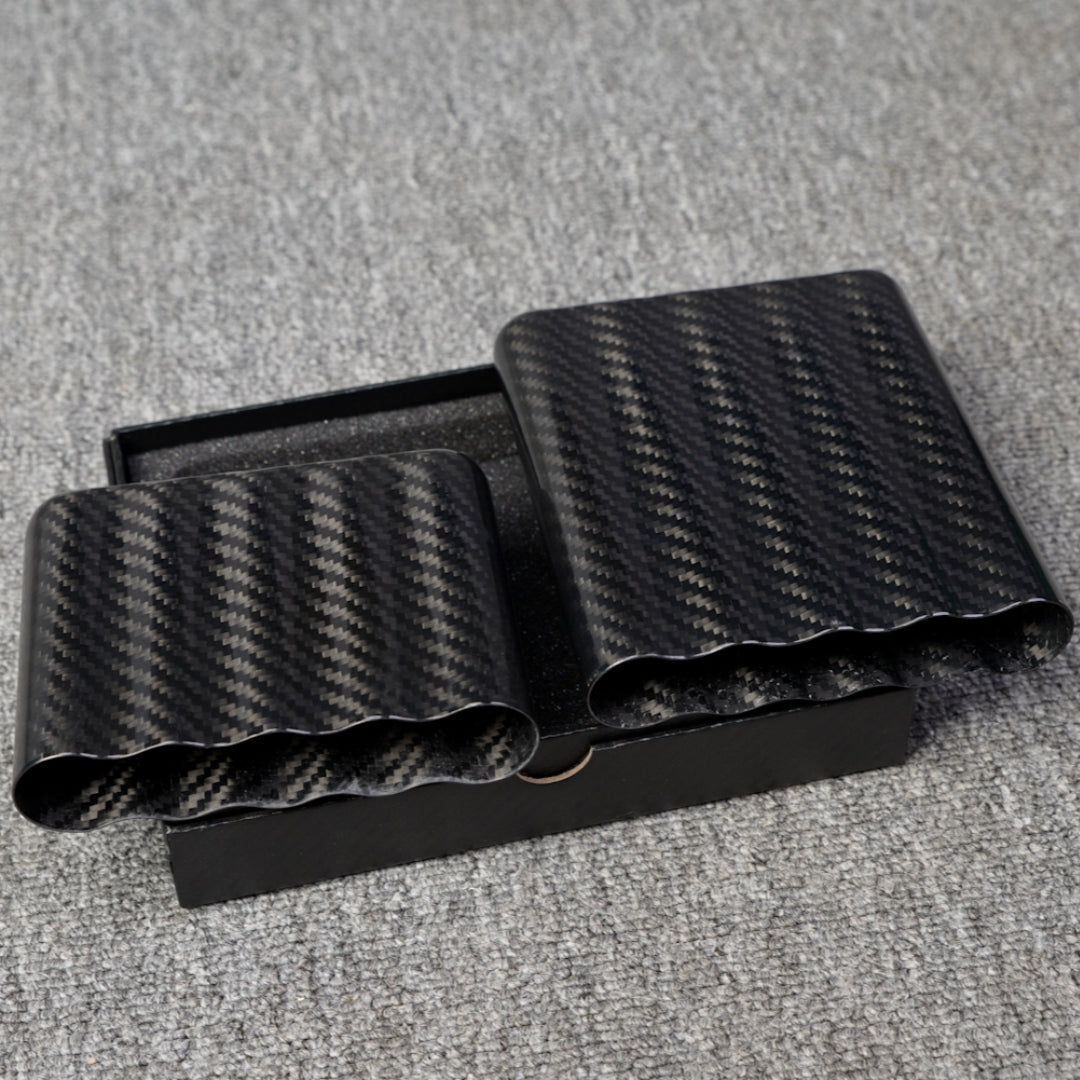Close-up of two carbon fiber cigar tubes (each capable of holding 5 cigars) with a distinct weave pattern, placed in an open black box on a gray fabric surface.