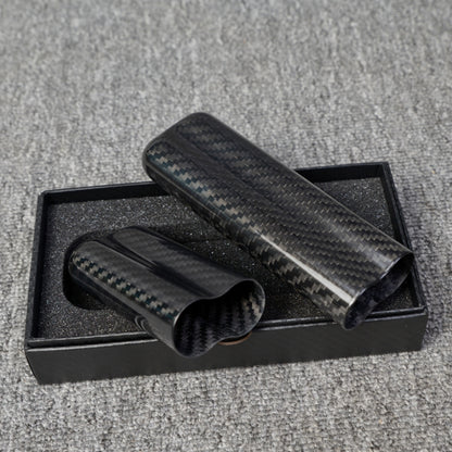 Close-up of two carbon fiber cigar tubes (capable of holding 2 cigars) with a distinct weave pattern, placed in a black box with foam lining on a gray fabric surface.