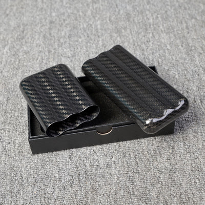 Close-up of carbon fiber cigar tubes with a capacity for 3 cigars, featuring a distinct weave pattern, placed in a black box on a gray fabric surface.