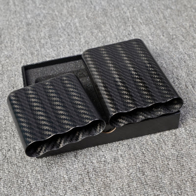 Close-up of carbon fiber cigar tubes with a capacity for 4 cigars, featuring a distinct woven pattern, placed in a black box on a gray fabric surface.