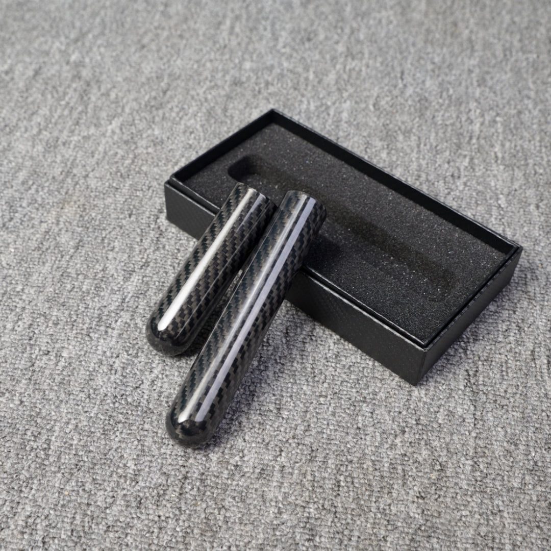 Close-up of two carbon fiber cigar tubes with single-cigar capacity, featuring a distinct woven pattern, placed near an open black box with foam lining on a gray fabric surface.