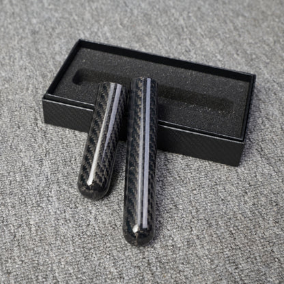 Close-up of two carbon fiber cigar tubes with single-cigar capacity, featuring a distinct woven pattern, placed next to an open black box with foam lining on a gray fabric surface.