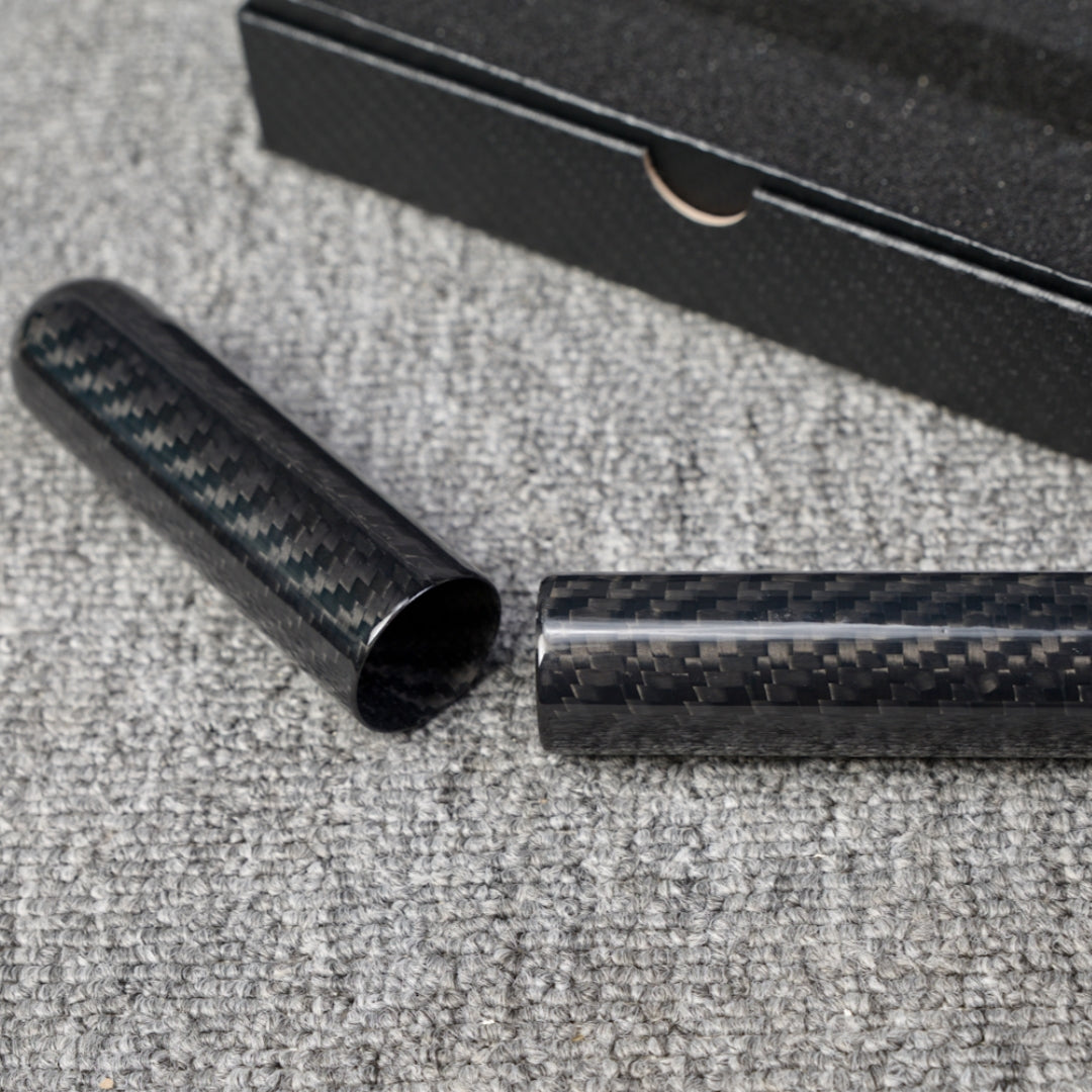 Close-up of a carbon fiber cigar tube (single-cigar capacity) with a distinct woven pattern, displayed in a disassembled state, placed on a gray fabric surface with a partial view of a black box in the background.