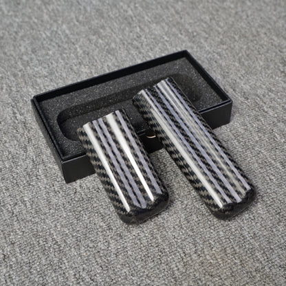 Close-up of two carbon fiber cigar tubes with a distinct striped carbon fiber pattern, placed near an open black box with foam lining on a gray fabric surface.
