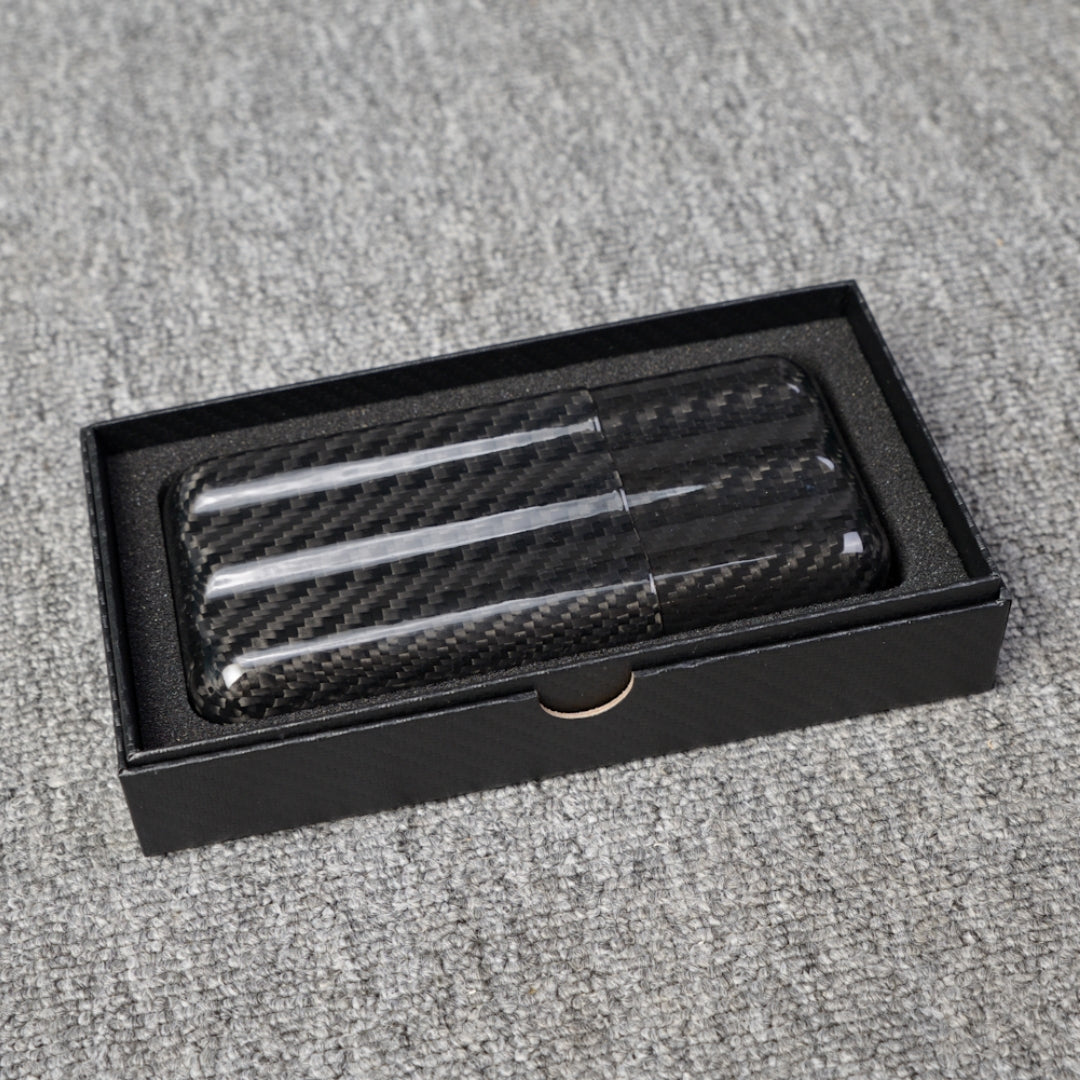 Close-up of a carbon fiber cigar tube with a capacity for 3 cigars, featuring a distinct woven pattern, placed in an open black box with foam lining on a gray fabric surface.