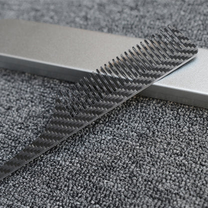 Close-up of a carbon fiber comb with a distinct woven pattern, placed next to a silver metal case on a dark gray textured fabric surface.