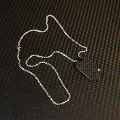 Close-up of a 3K carbon fiber dog tag pendant with a checkered pattern and a small diamond accent, paired with a silver-tone chain, displayed on a carbon fiber textured background.