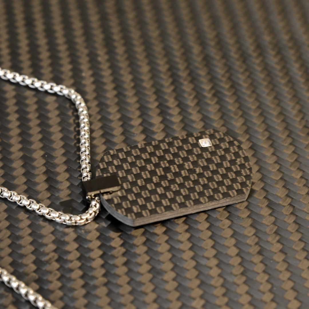 Close-up of a 3K carbon fiber dog tag pendant with a checkered pattern and a small diamond accent, paired with a silver-tone chain, displayed on a carbon fiber textured background.