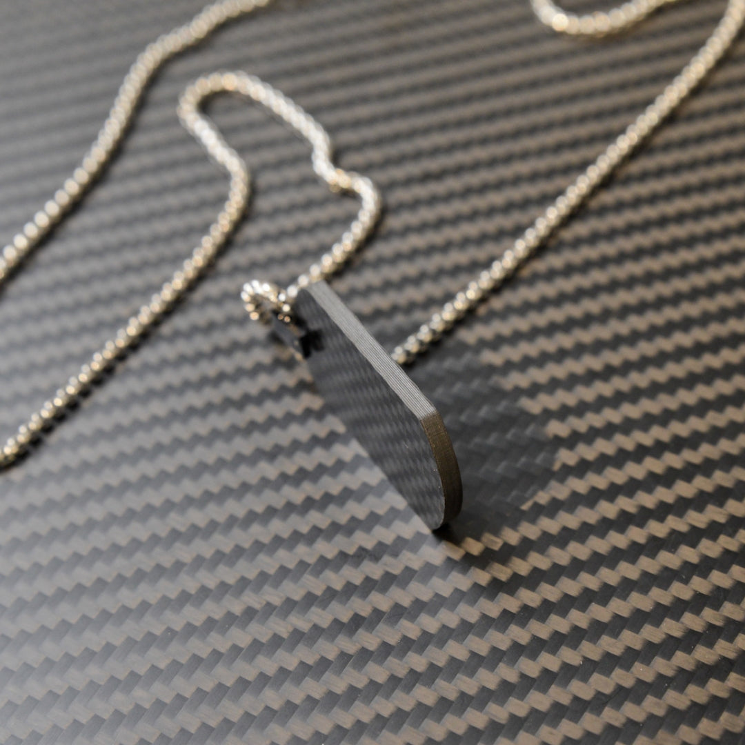 Close-up of a 3K carbon fiber dog tag pendant with a sleek, textured surface, paired with a silver ball chain, displayed on a matching carbon fiber patterned background.