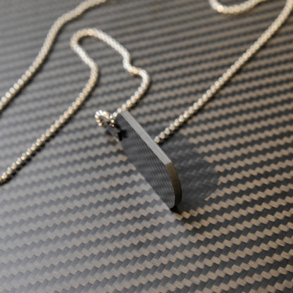 Close-up of a 3K carbon fiber dog tag pendant with a sleek, textured surface, paired with a silver ball chain, displayed on a matching carbon fiber patterned background.