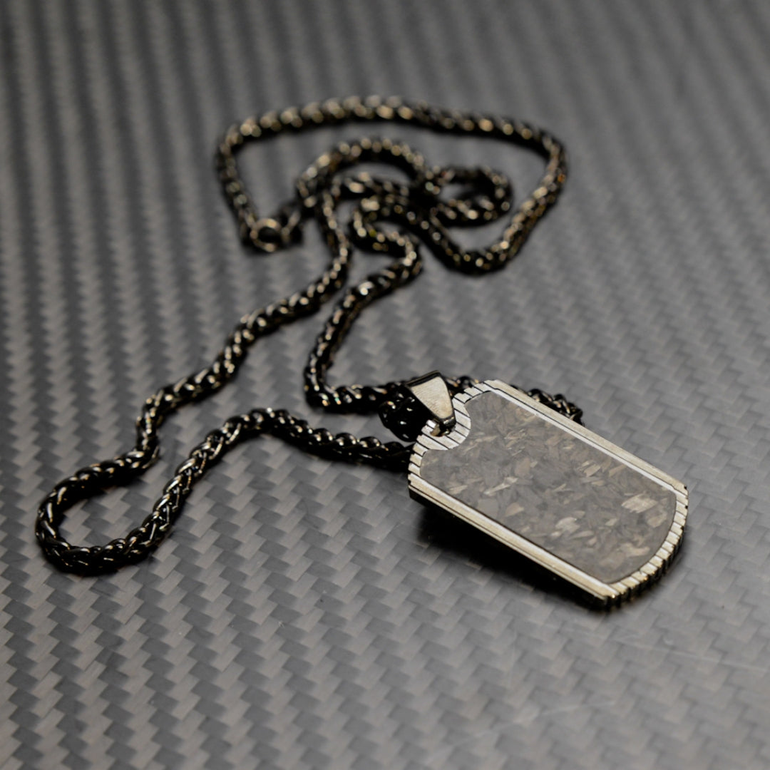 Close-up of a 3K carbon fiber dog tag pendant with metallic detailing, paired with a black-and-gold-toned chain, displayed on a carbon fiber textured background.