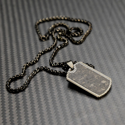 Close-up of a 3K carbon fiber dog tag pendant with metallic detailing, paired with a black-and-gold-toned chain, displayed on a carbon fiber textured background.