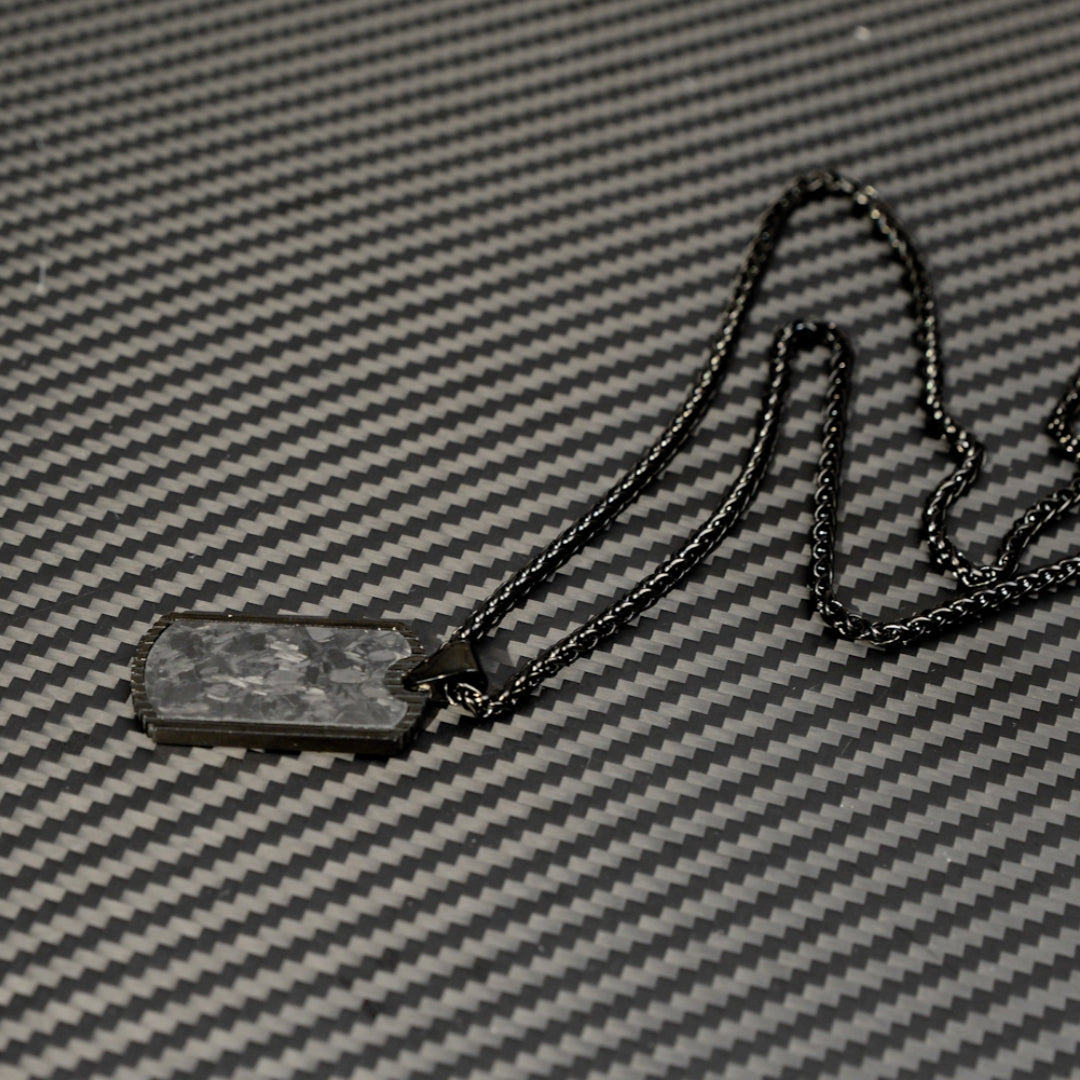 Close-up of a 3K carbon fiber dog tag pendant with a textured surface, paired with a black-toned chain, displayed on a matching carbon fiber patterned background.