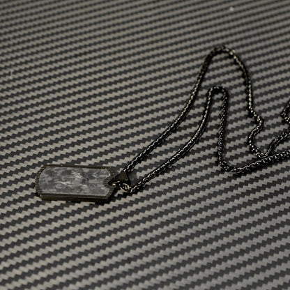 Close-up of a 3K carbon fiber dog tag pendant with a textured surface, paired with a black-toned chain, displayed on a matching carbon fiber patterned background.