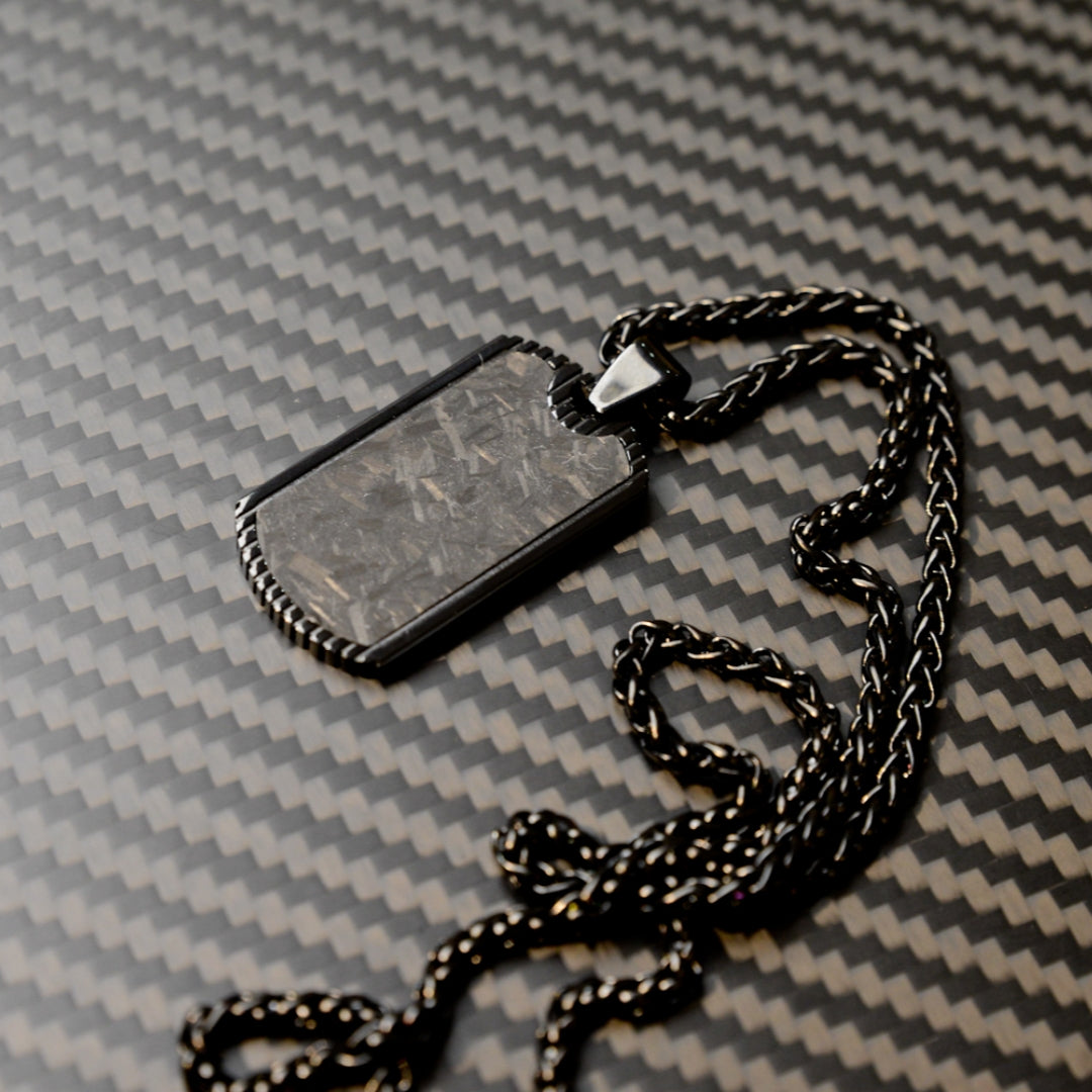 Close-up of a 3K carbon fiber dog tag pendant with a textured surface, paired with a black-toned chain, displayed on a carbon fiber patterned background.