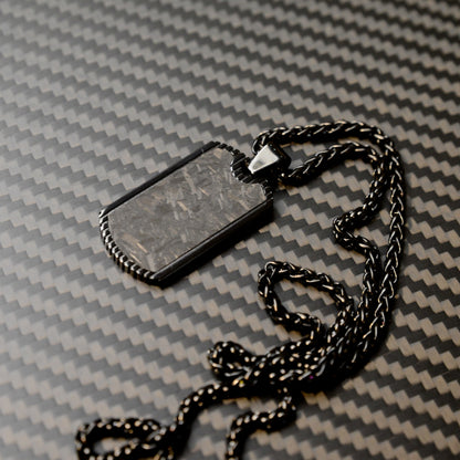 Close-up of a 3K carbon fiber dog tag pendant with a textured surface, paired with a black-toned chain, displayed on a carbon fiber patterned background.