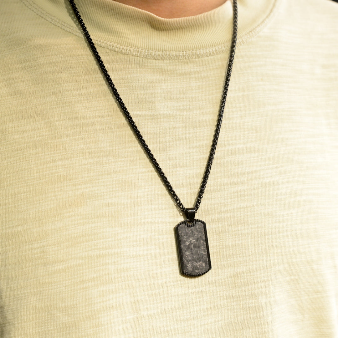 Close-up of a 3K carbon fiber dog tag pendant with a textured surface, paired with a black chain, worn on a light beige T-shirt.