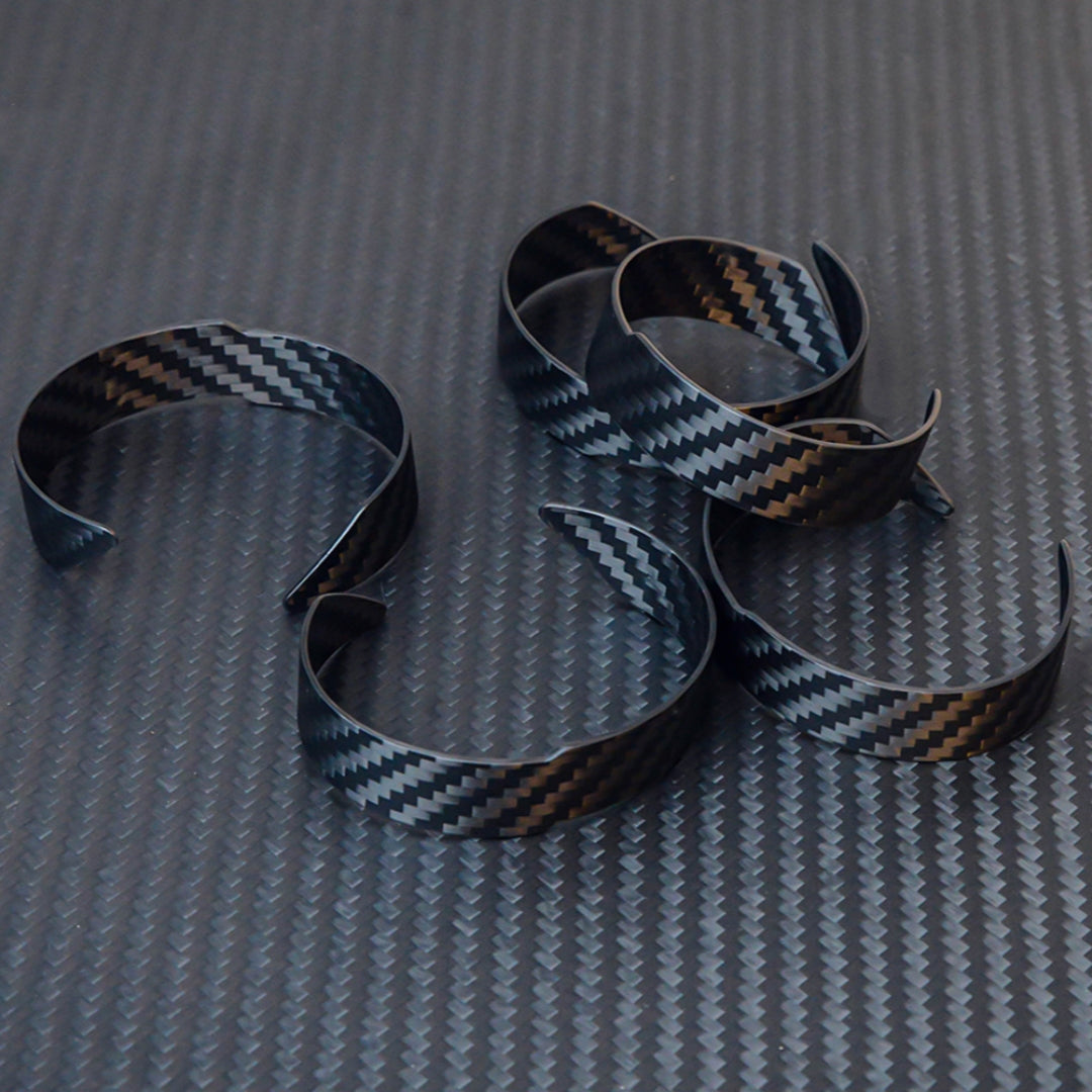 Several 3K carbon fiber cuff bracelets with a distinct chevron weave pattern, placed on a dark carbon fiber textured surface.
