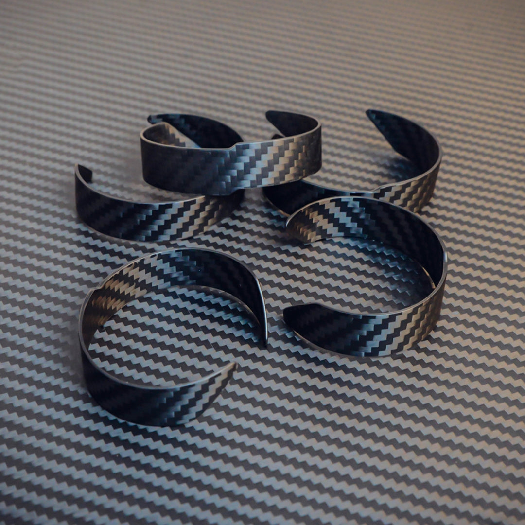Several 3K carbon fiber cuff bracelets with a distinct weave pattern, arranged on a matching carbon fiber textured surface.