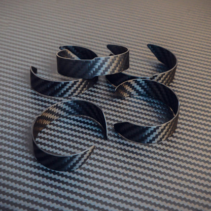 Several 3K carbon fiber cuff bracelets with a distinct weave pattern, arranged on a matching carbon fiber textured surface.