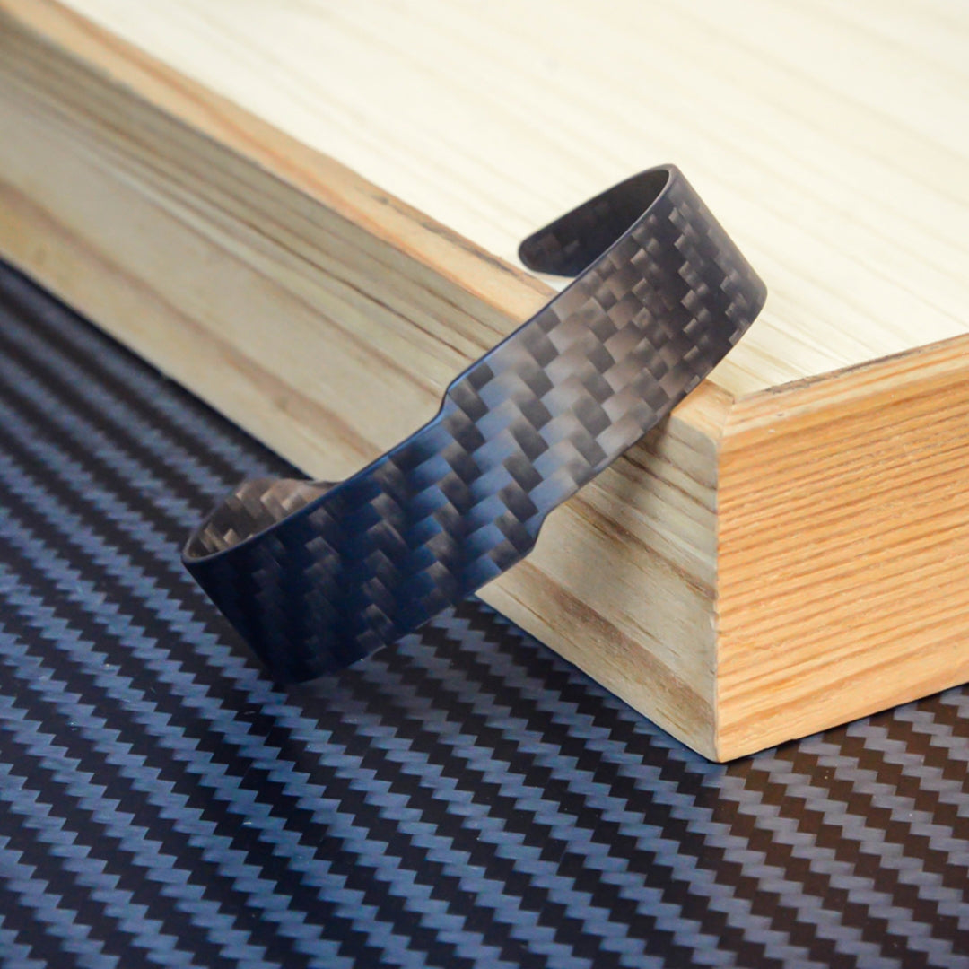 A 3K carbon fiber cuff bracelet with a distinct weave pattern, resting on a wooden block against a carbon fiber textured background.