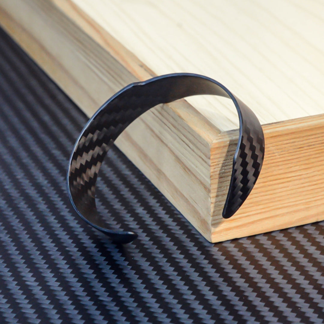 A 3K carbon fiber cuff bracelet with a distinct weave pattern, resting on a wooden block against a carbon fiber textured background.