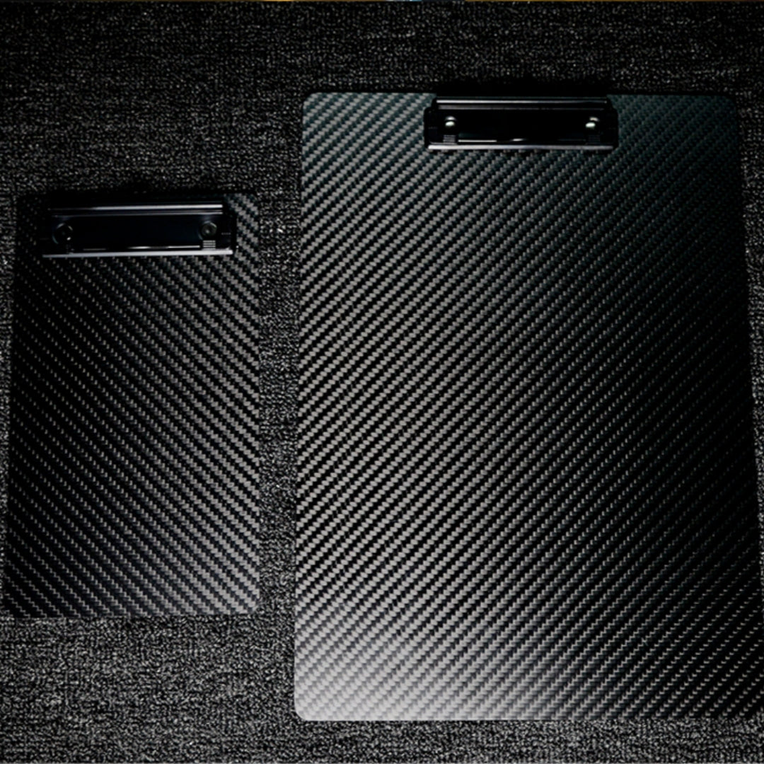 Close-up detail of two 3K carbon fiber clipboards featuring a distinctive woven pattern and black metal clips, placed on a textured dark surface.