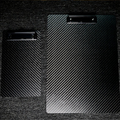 Close-up detail of two 3K carbon fiber clipboards featuring a distinctive woven pattern and black metal clips, placed on a textured dark surface.