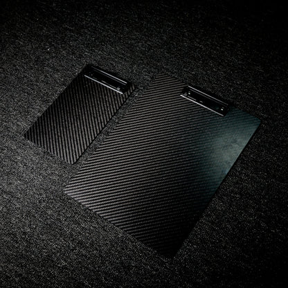 Close-up detail of two 3K carbon fiber clipboards with a distinct woven pattern and black metal clips, placed on a dark textured surface.