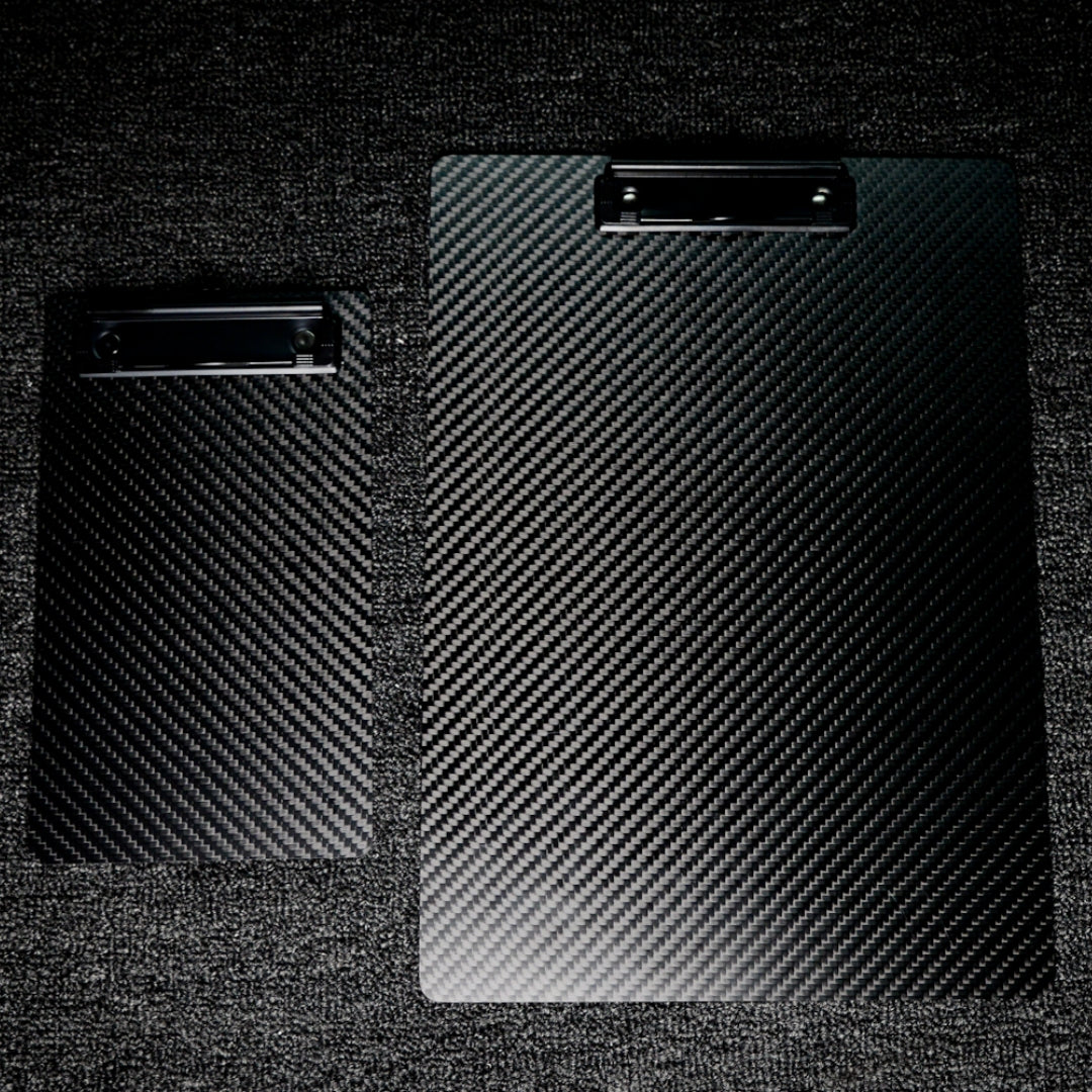 Close-up detail of two 3K carbon fiber clipboards featuring a distinctive woven pattern and black metal clips, positioned on a dark textured surface.